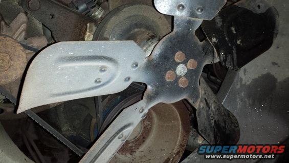 Close-up of a vehicle's engine fan and belts. The fan has four blades and a central hub with five circular rivets