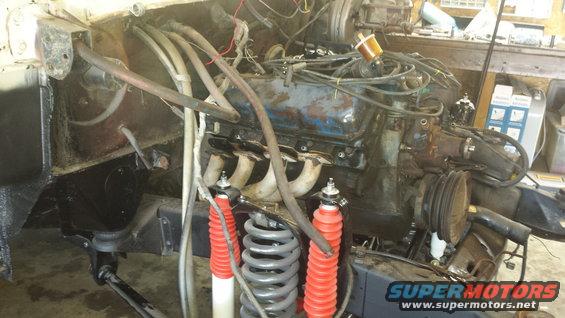 Engine bay of a vehicle under repair, showing a V8 engine, exhaust manifolds, red shock absorbers, and coil springs
