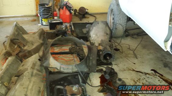 Disassembled vehicle parts including a transmission, fan shroud, and chassis components scattered on a concrete floor