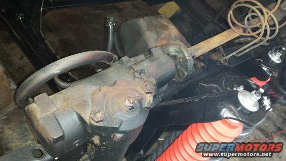 Close-up of a rusty steering gear box with hoses and black painted suspension parts, including a red coil spring