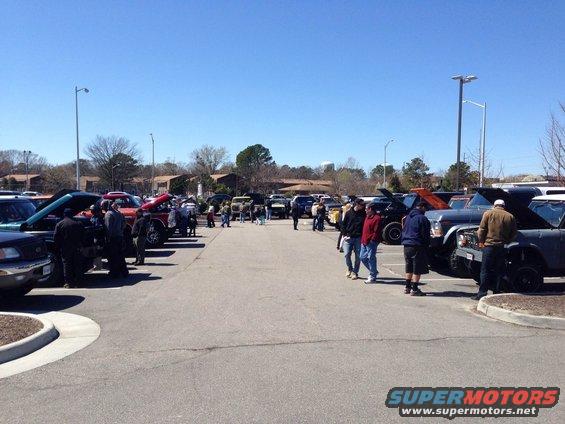 Parking lot filled with various vehicles and people gathered around them under a clear blue sky