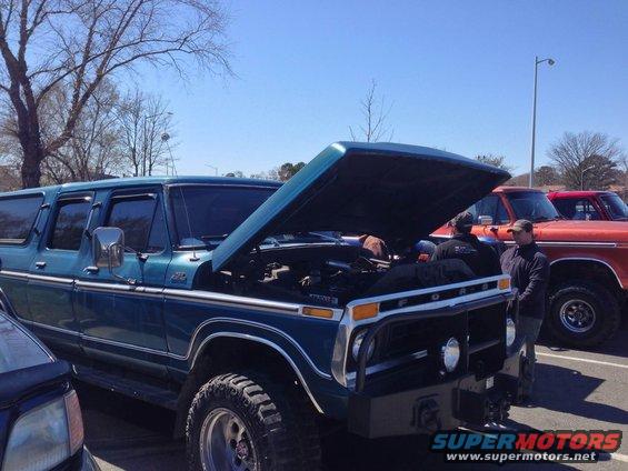 Blue Ford Bronco SUV, hood open, showing engine. Another orange Ford truck is parked behind it