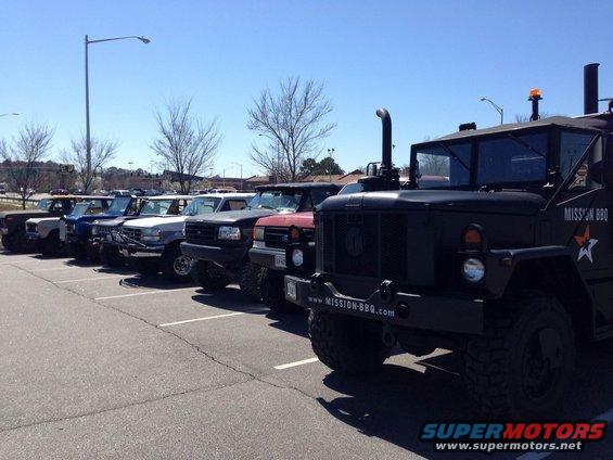 Several trucks and SUVs parked in a lot under a blue sky. A large black military-style truck is in the foreground