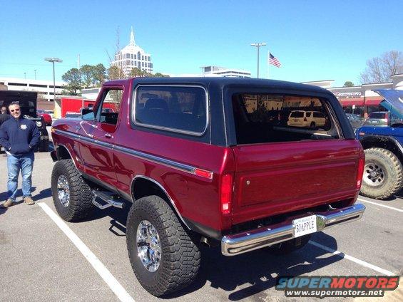 Red 1978 Ford Bronco with lifted suspension and black top, parked next to a man. Another vehicle is visible to the right