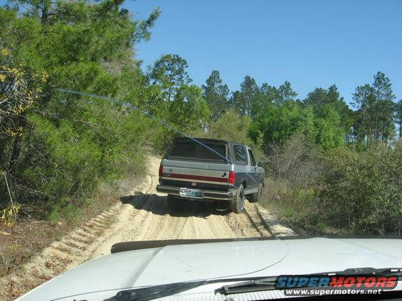 1994 Ford Bronco driving on a sandy dirt road through trees. Another vehicle is visible in the foreground