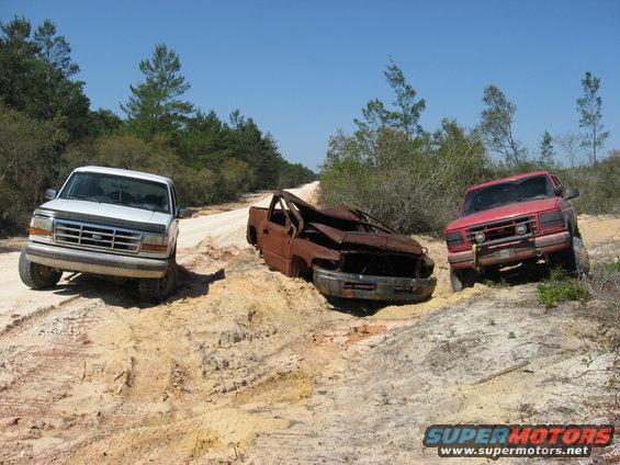 White truck, rusty wrecked truck, and red truck stuck on a dirt road