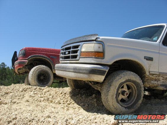 White Ford Bronco with a black hood deflector and a red Ford Bronco behind it, both on a dirt mound
