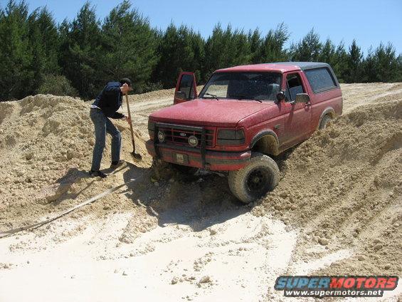 Red Ford Bronco stuck in sand, driver digging with shovel. Front grille and headlights visible