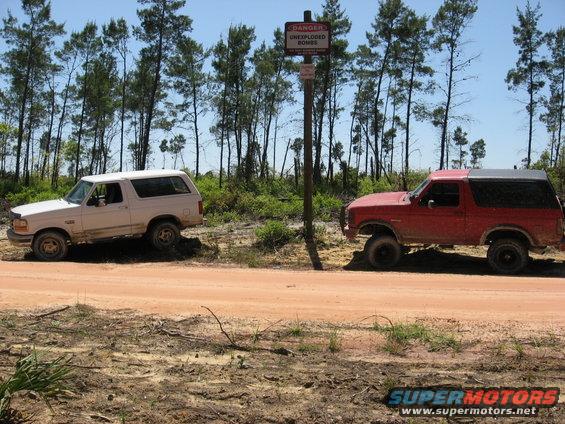 Two Ford Broncos parked on a dirt road near a sign warning of unexploded bombs