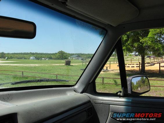 Interior view from driver's seat looking out windshield at a rural scene with buildings and trees. Side mirror reflects trees