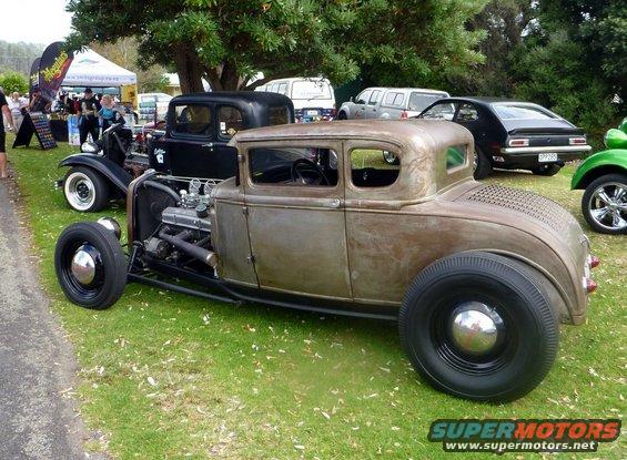 Rusty hot rod coupe with exposed engine and large rear tires parked on grass