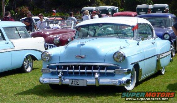 Light blue 1953 Chevrolet Bel Air sedan with a white roof, chrome grille, and "ACE 1" license plate. Other classic cars are visible in the background