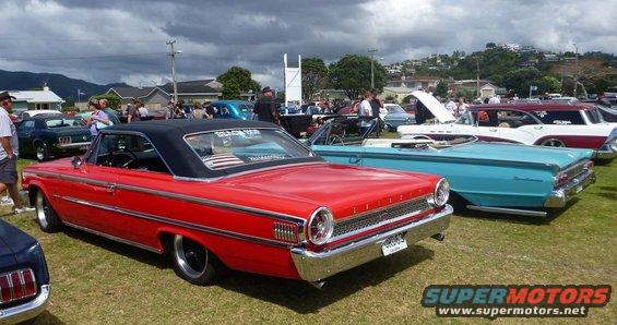 Red classic car with a black top parked on grass at an outdoor car show. Other classic cars are visible in the background