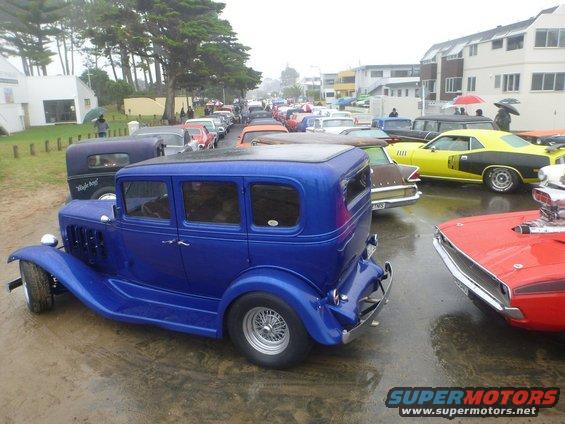 Blue hot rod sedan parked on wet pavement, other classic cars in background