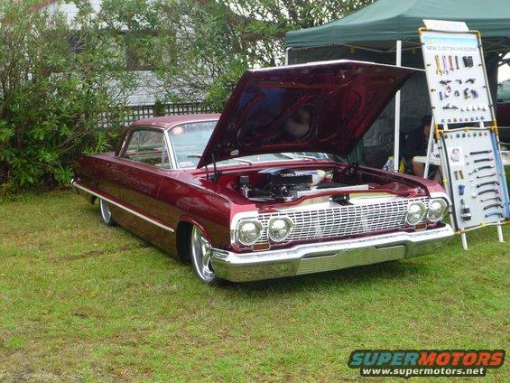 maroon 1963 Chevrolet Impala with hood open, showing engine. Display stand with parts nearby