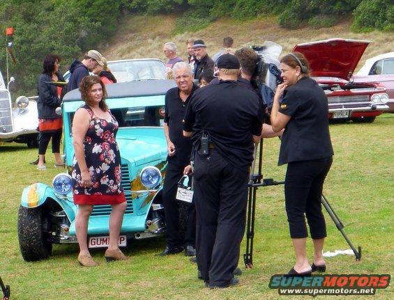 Woman in floral dress stands by a light blue hot rod with a film crew nearby. Other vintage cars are in the background