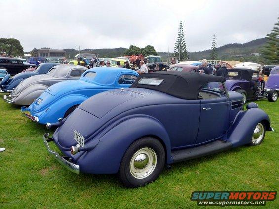 Dark blue convertible hot rod with black top, parked on grass with other classic cars