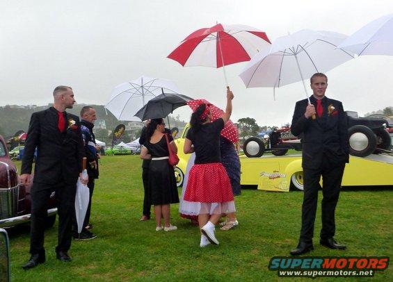 People in formal wear and 1950s style dresses holding umbrellas at an outdoor car show event