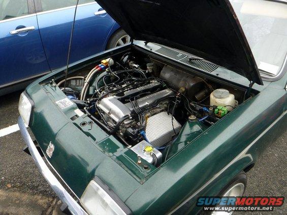 Green car engine bay with a V8 engine, chrome accents, and visible hoses. A blue car is parked beside it