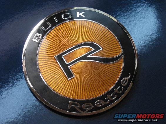 Close-up of a circular Buick Reatta emblem with a gold sunburst background and a black 'R' logo