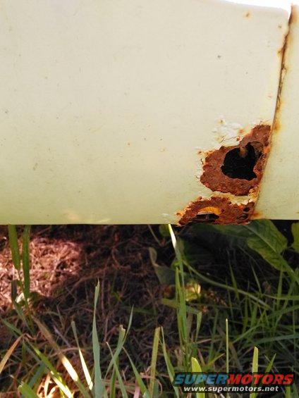 Close-up of a rusted-out section on a cream-colored vehicle panel, with grass and dirt visible below