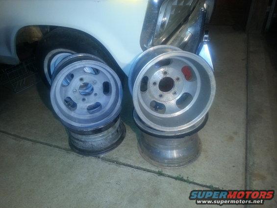 Two aftermarket wheels stacked on top of other wheels, with a vehicle fender and tire in the background