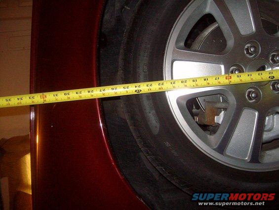Measuring tape across a car's wheel well, showing the tire and silver alloy wheel