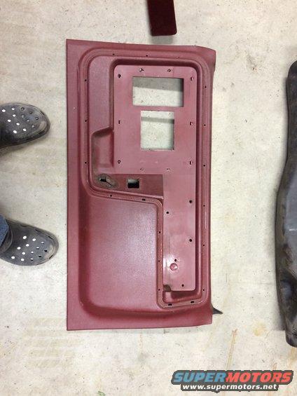 Red car door panel with cutouts for window and speaker, lying on concrete floor next to a dark metal part
