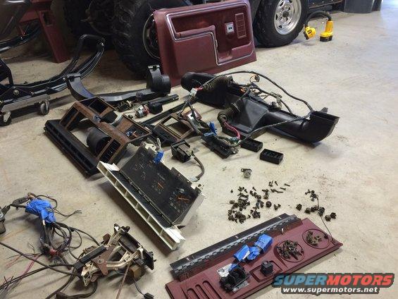 Vehicle interior parts, including dashboard components, wiring harnesses, and a door panel, are spread out on a concrete floor