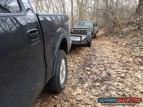 Black Ford Bronco on a trailer, towed by a black pickup truck on a dirt road