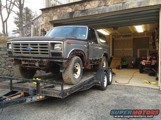 Gray Ford Bronco on a trailer in front of a garage. Some rust visible on the body