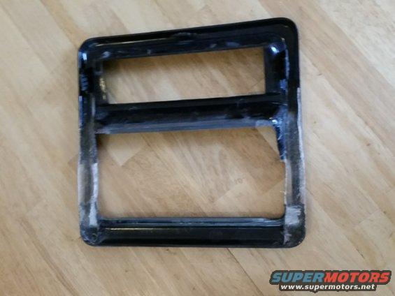 Black plastic trim piece with two rectangular openings, sitting on a wood floor. Supermotors.net logo visible