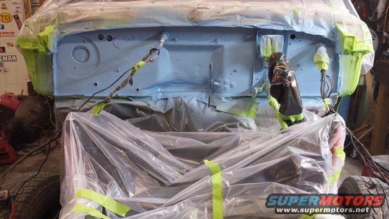 Vehicle firewall covered in blue primer and plastic sheeting, with green tape on edges