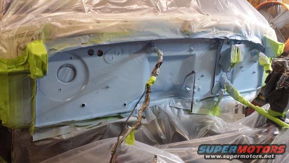Engine bay firewall covered in light blue primer, masked with green tape, and draped in plastic sheeting