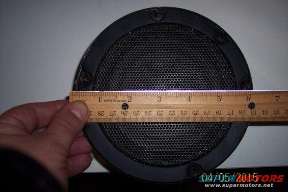Round car speaker with black grille and frame, measured with a wooden ruler