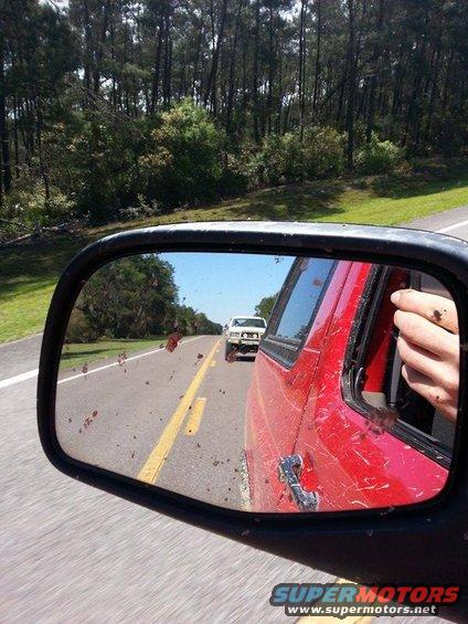 Side mirror view of a white pickup truck driving on a road, seen from a red vehicle