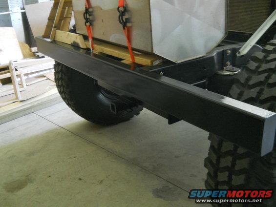 Black aftermarket rear bumper on a vehicle with large off-road tires. A cargo box is strapped to the frame