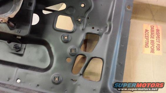 Close-up of the inside of a car door panel, showing metal bracing and holes. A yellow sticker reads 'INSPECT FOR DAMAGE BEFORE ACCEPTING'