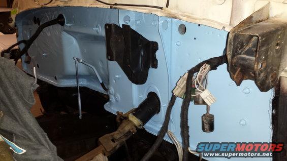 Close-up of a vehicle's firewall with light blue paint and a black wiring harness. A metal bracket is attached to the right