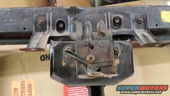 Close-up of a vehicle's hood latch assembly. Some text is visible on cardboard in the background