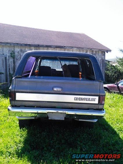 Rear view of a dark blue Chevrolet K5 Blazer parked on grass in front of a barn. Another car is visible to the right