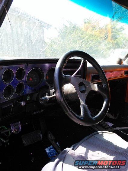 Interior shot of a vehicle dashboard with a steering wheel, gauges, and pedals visible. A person's leg is in the foreground