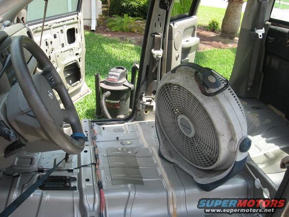 Interior of a vehicle with doors removed, showing a fan and shop vac