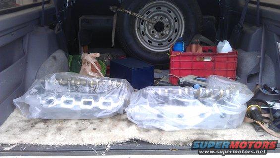 Two engine camshafts and exhaust manifolds wrapped in plastic, stored in the cargo area of a vehicle with a spare tire and red crate