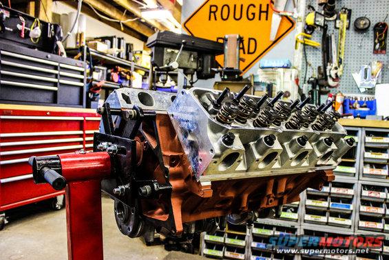 V8 engine on a red stand in a garage with tool chests and a 'Rough Road' sign