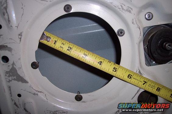 Measuring a door speaker mounting hole with a yellow tape measure. The hole is approximately 6 inches in diameter