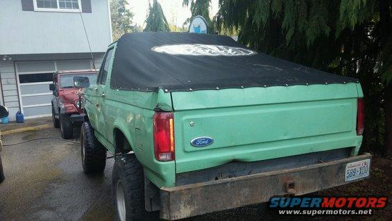 Green 1990 Ford Bronco with black soft top, parked next to a red Jeep