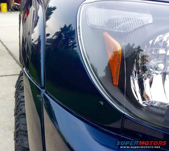 Close-up of dark blue SUV fender and headlight assembly, showing tire and part of the grille. Watermark reads SUPERMOTORS.NET