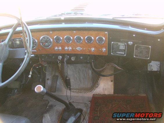 Truck interior with wood-grain dash, gauges, steering wheel, and floor shifter. Small rug on floor