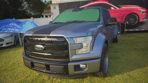 Gray Ford F-150 pickup truck with black racing stripes, parked on grass. A red Ford Mustang is on a trailer behind it